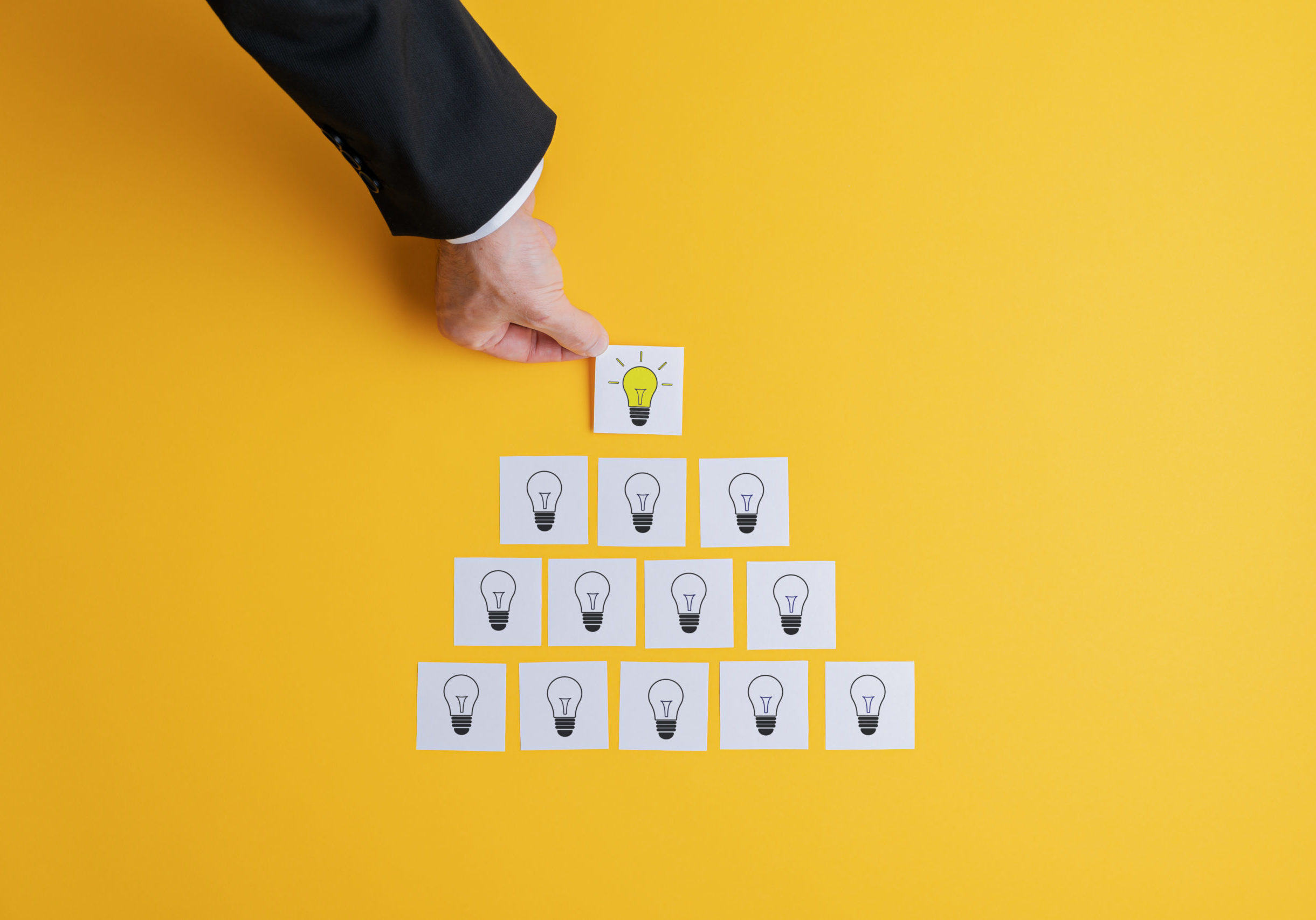 Businessman making pyramid shape of post it papers with light bulb drawn on them. Over yellow background.
